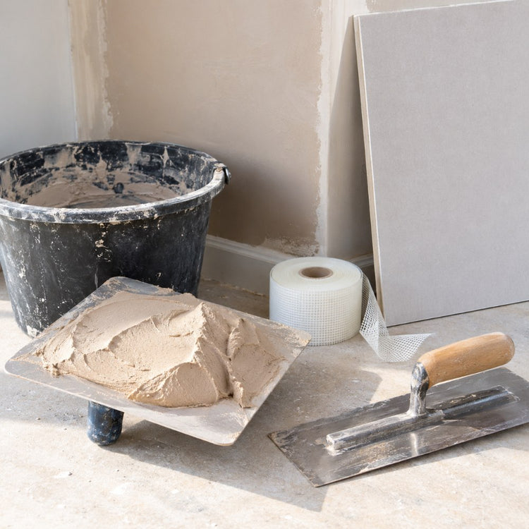 A plastering trowel and hawk with fresh plaster, a bucket, jointing tape and a sheet of plasterboard against a wall